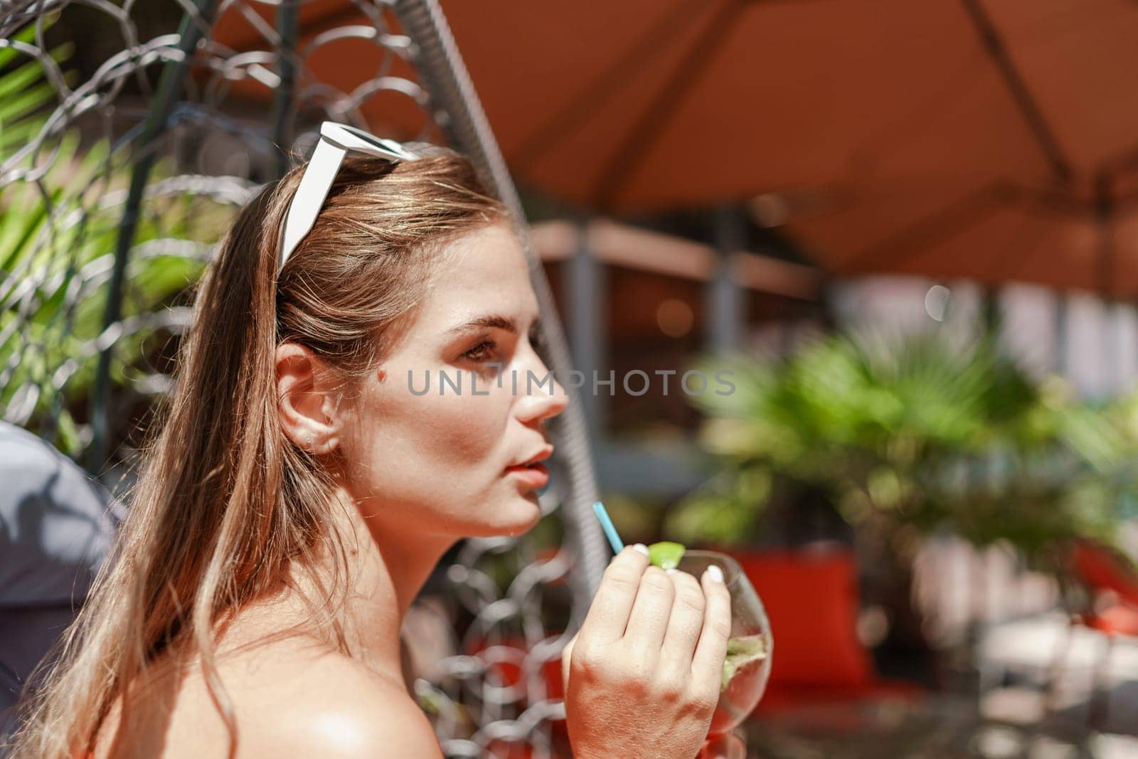 A woman in an orange dress is sitting in a chair with a drink in front of her. She is looking at the camera and she is enjoying her drink. The scene is set in a patio or outdoor area