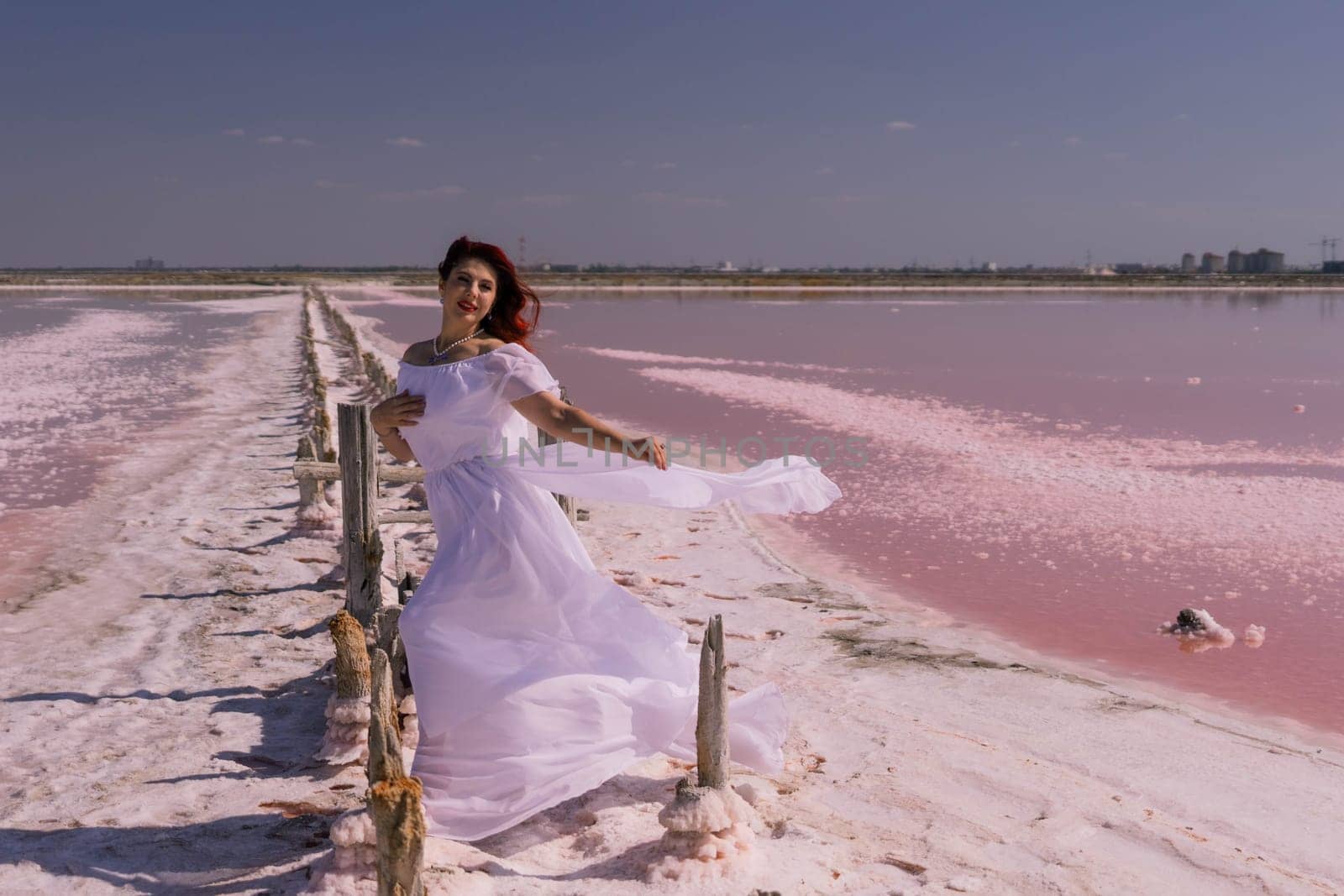 A woman in a white dress stands on a wooden walkway over a pink salt lake, her dress billowing in the wind.