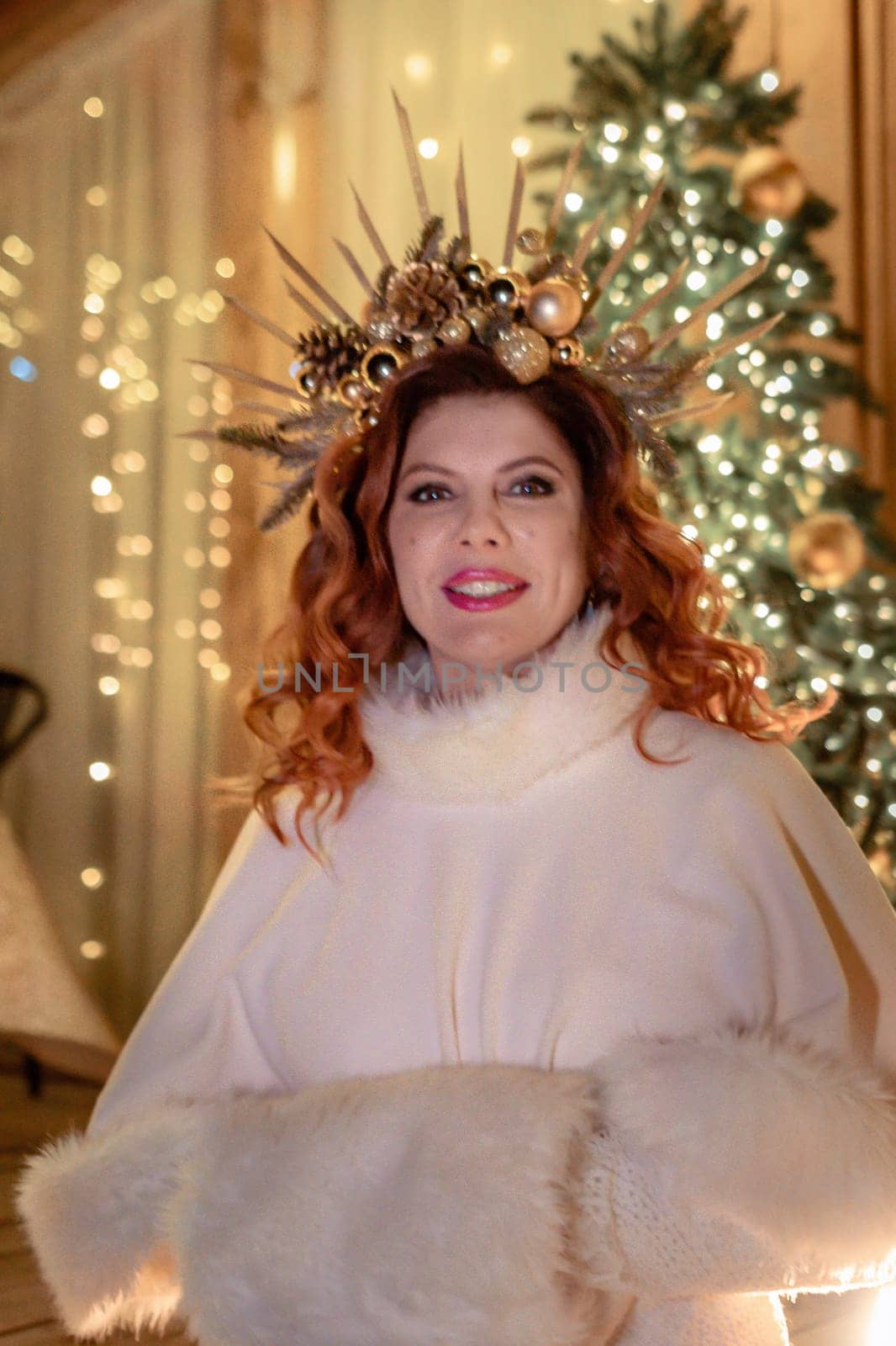 A woman with red hair and a white fur coat poses in front of a Christmas tree, wearing a golden crown of branches and ornaments.
