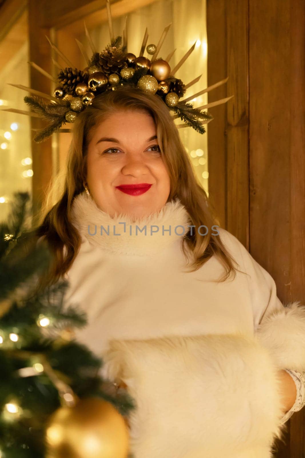 A woman with a festive wreath in her hair smiles while standing beside a Christmas tree.