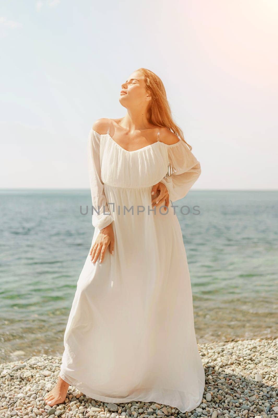 woman white dress stands on a beach, looking out at the ocean. She is in a contemplative or reflective mood, as she raises her hands above her head. Concept of peace and tranquility