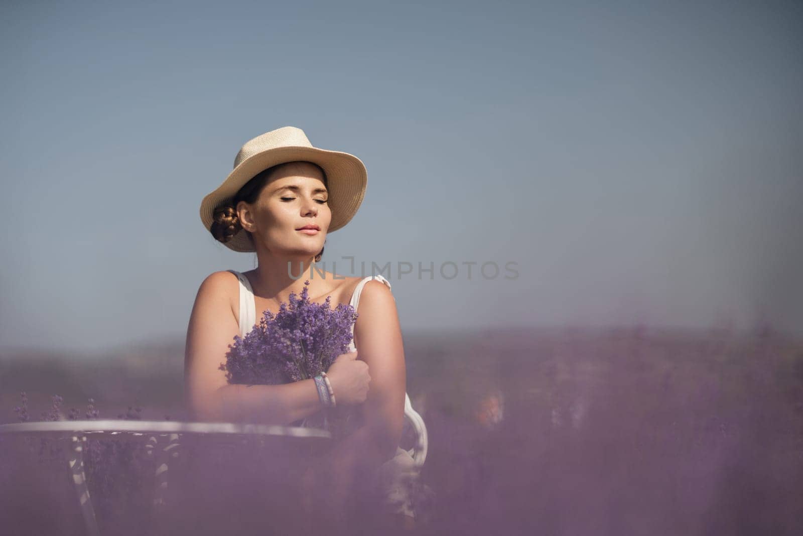 A woman is sitting in a field of lavender flowers and wearing a straw hat. She is smiling and holding a bouquet of flowers. Scene is peaceful and serene, as the woman is surrounded by the beauty of nature.