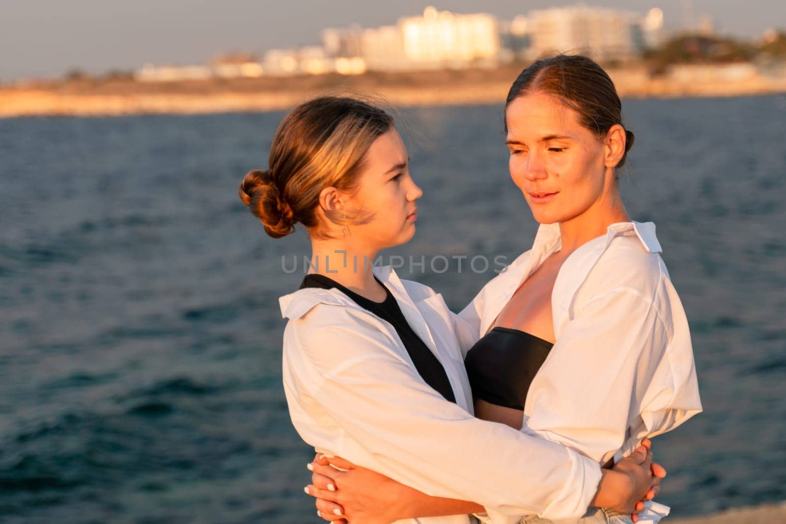 Two women, one in a white shirt and black tank top, the other in a white shirt, embrace by a calm ocean with a cityscape in the background.