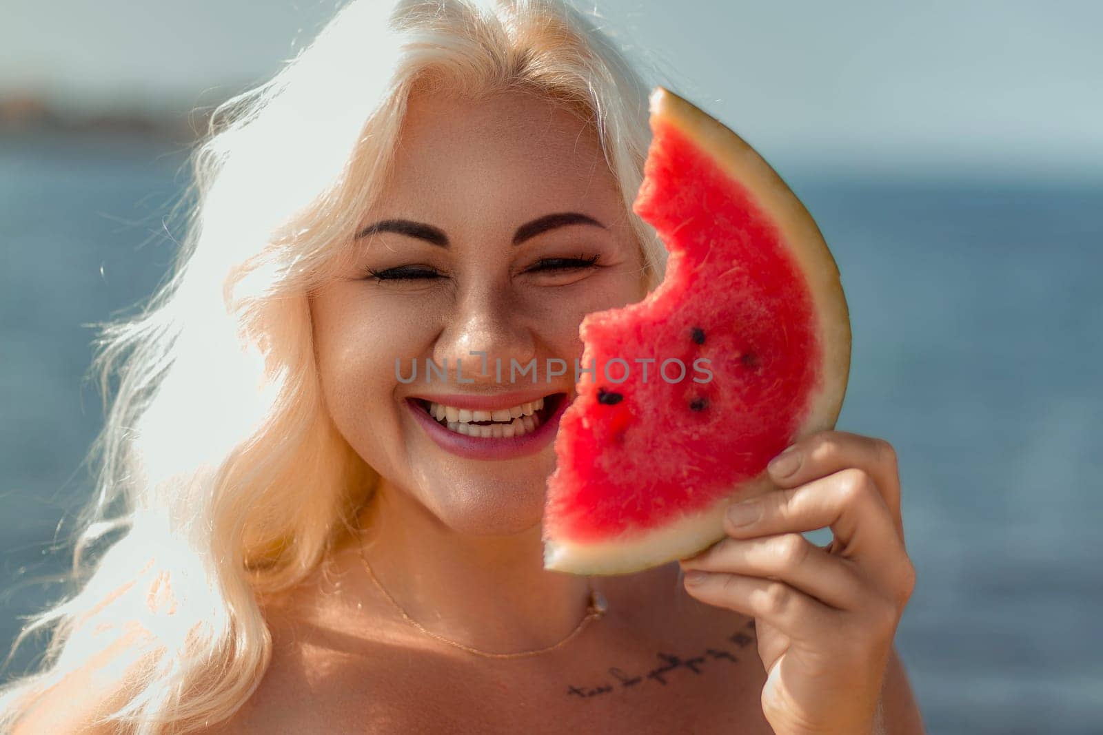 Smiling blonde woman holds a bitten watermelon slice, partially obscuring her face. Beach background visible.