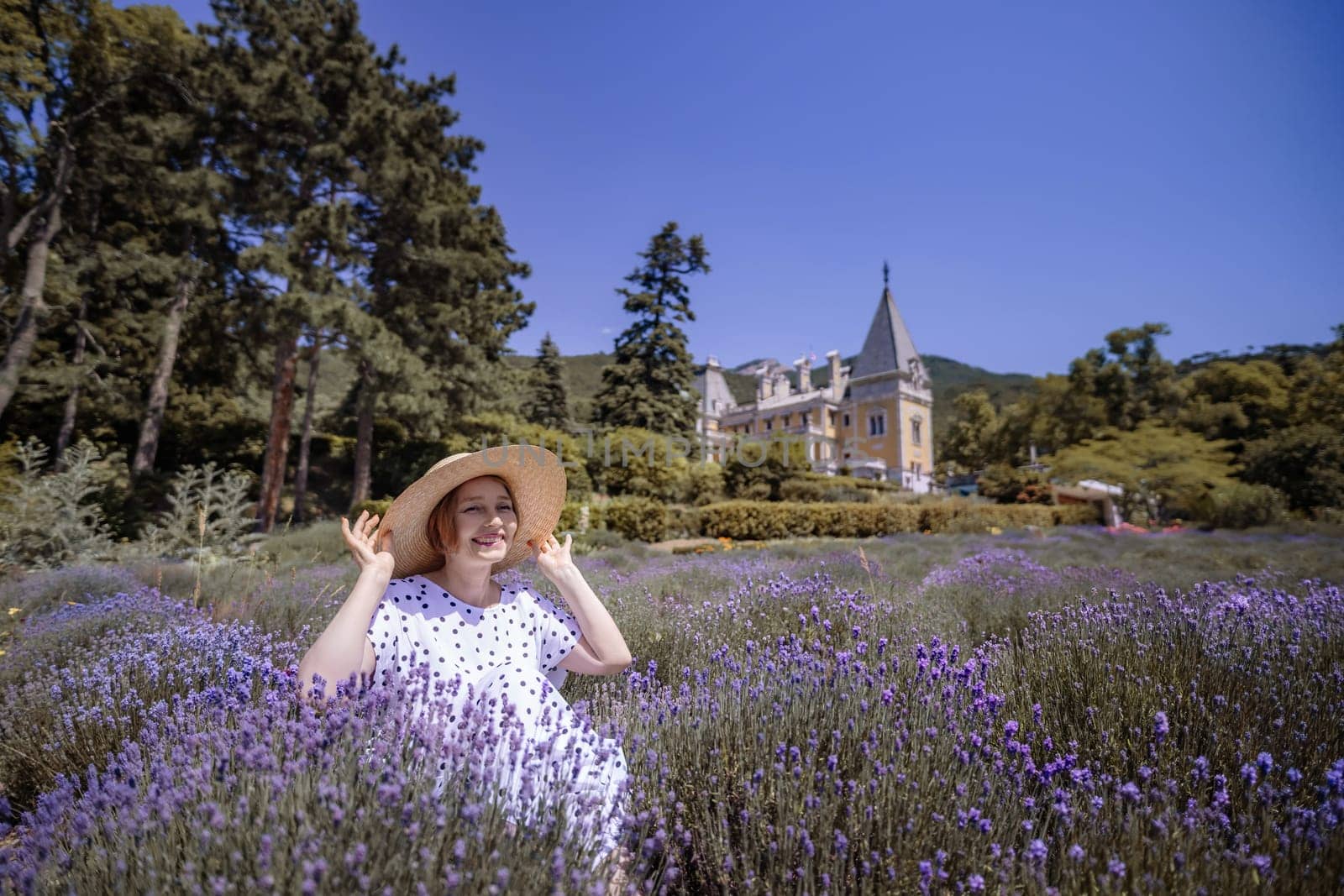 A woman is sitting in a field of purple flowers. She is wearing a straw hat and a white dress. The field is full of lavender, and the sky is clear and blue. The scene is peaceful and serene