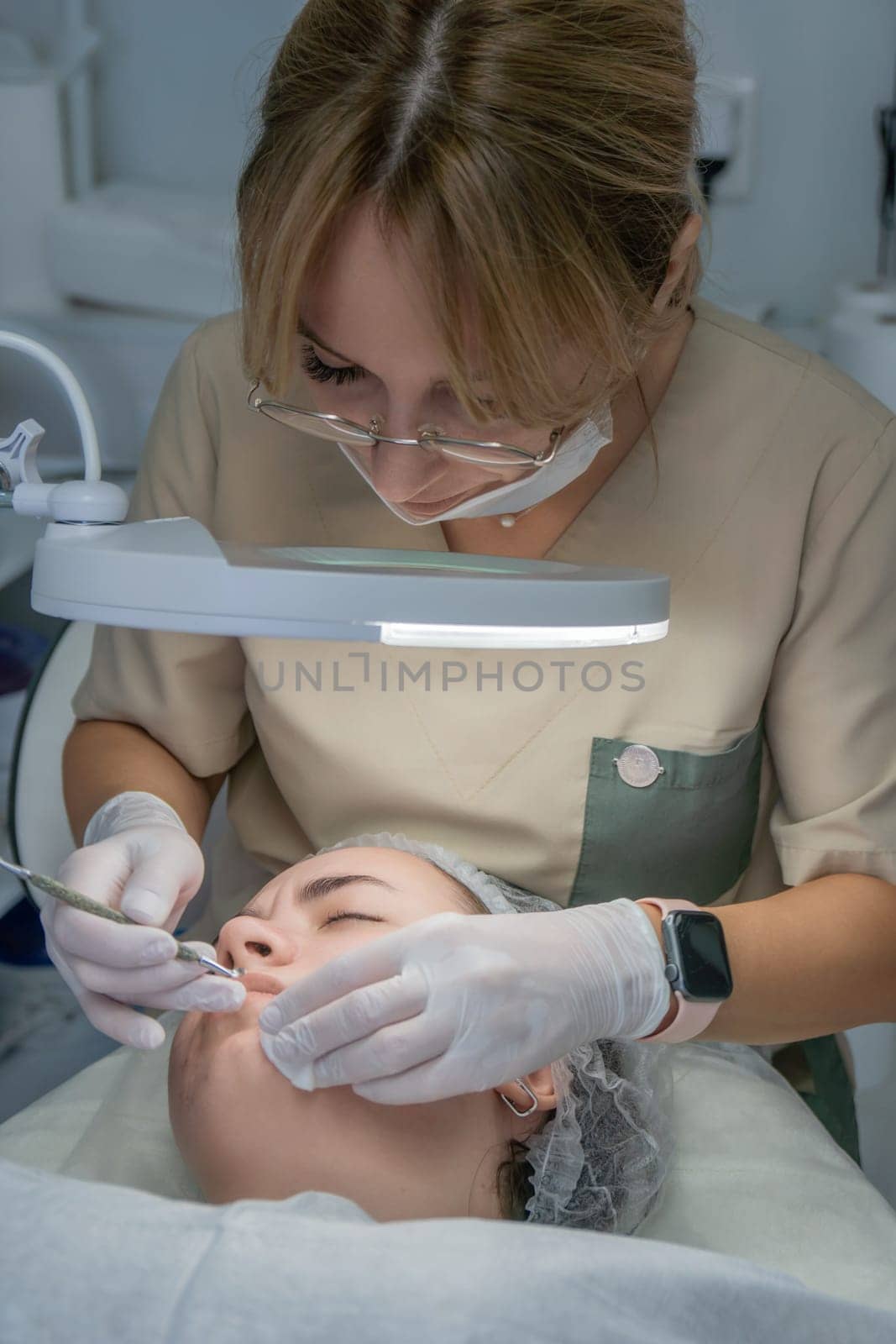 A beautician, wearing gloves and a mask, meticulously cleans a client's face using a small tool under a bright lamp. Facial treatment in progress.
