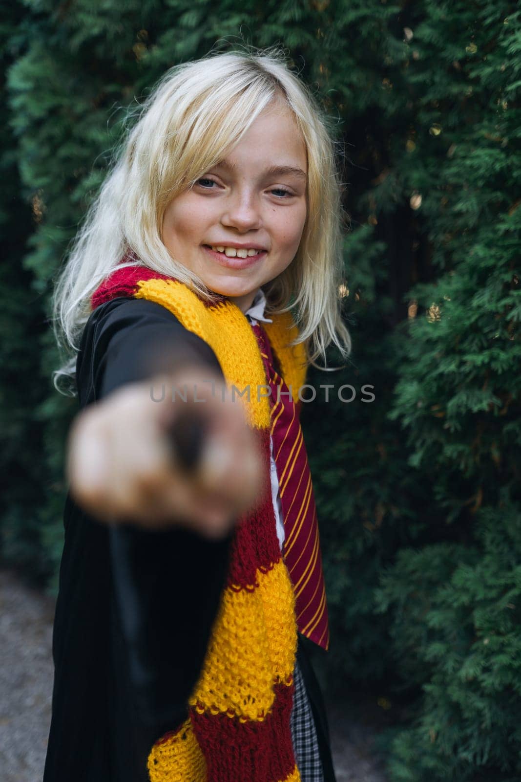 A young girl dressed in a Hogwarts uniform holds a wand out towards the camera, a mischievous smile on her face. She stands in front of a backdrop of evergreen trees.