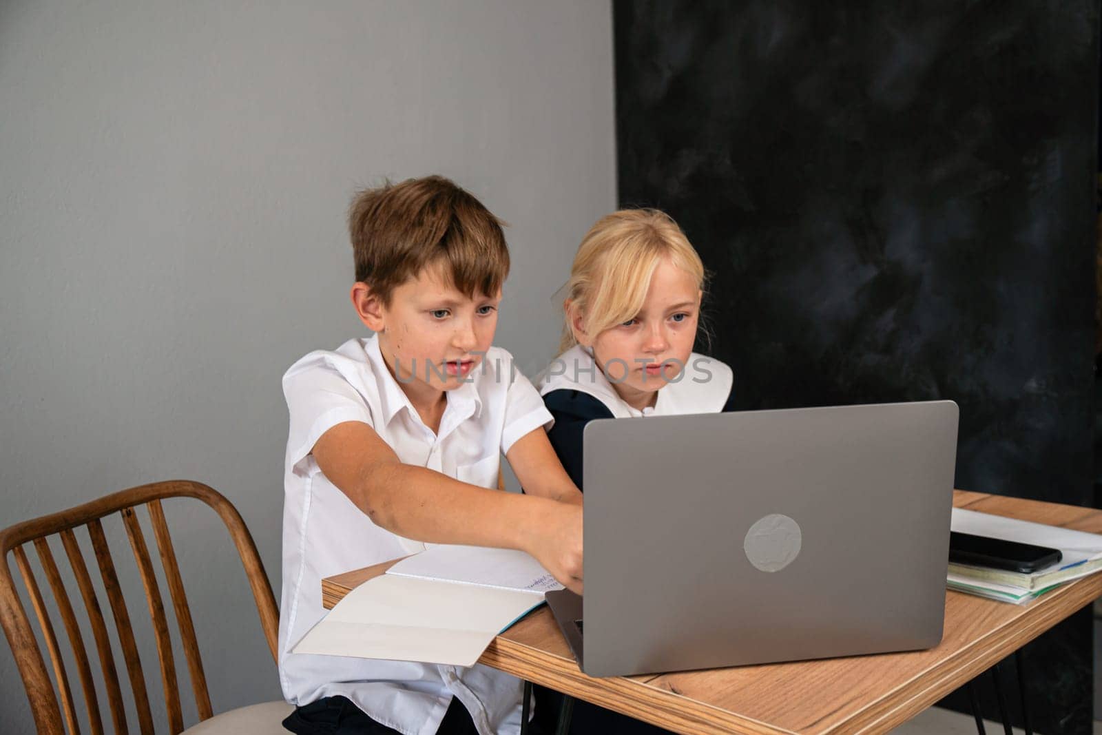 Two children, a boy and a girl, sit at a table and look at a laptop. The boy points at the screen while the girl watches.
