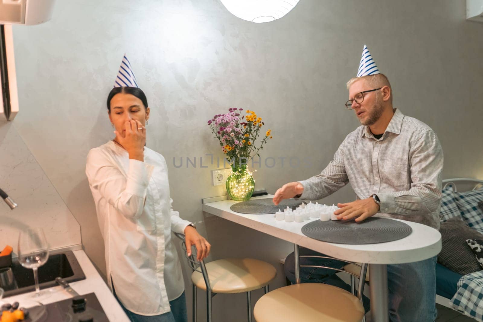 Here's a caption for the image: A man and woman, wearing party hats, sit at a small kitchen table. One pinches their nose, the other looks down at some scattered paper. Flowers are in a vase nearby.