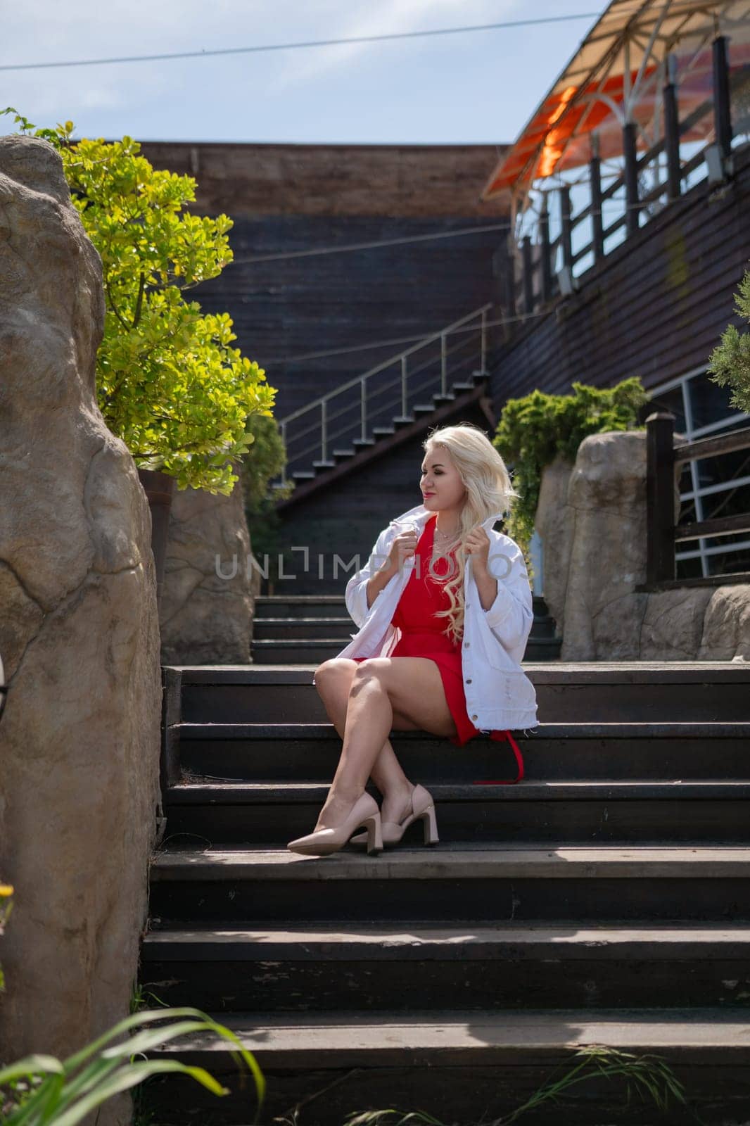 Blonde woman in red dress and white blazer sits on outdoor stairs. She's wearing heels with green foliage and a building in the background.