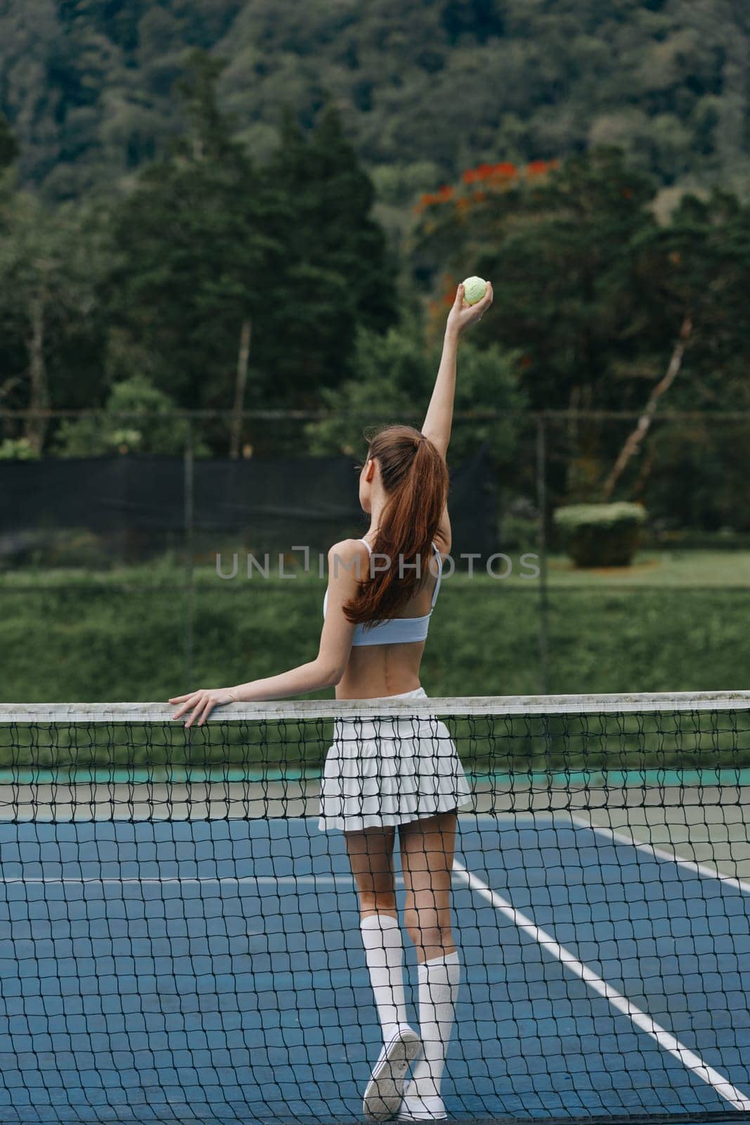 Energetic young female tennis player in a sporty outfit serving a ball on a sunny outdoor court, surrounded by lush greenery, portraying focus and athleticism