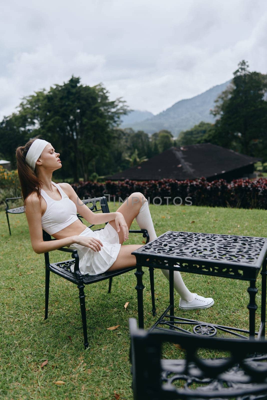 A young fitness model in a stylish white outfit enjoys a peaceful moment in a garden setting, combining elegance with an active lifestyle, surrounded by lush greenery and soft clouds