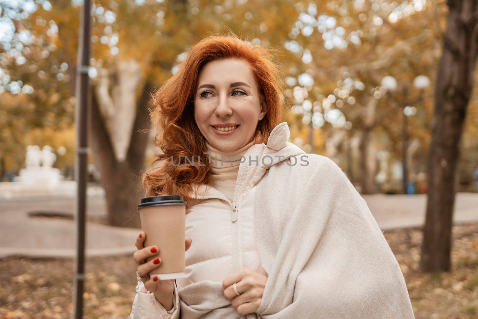 Redhead woman smiles, wrapped in a cream-colored shawl, holding a coffee cup outdoors in an autumnal park. Leaves are golden.