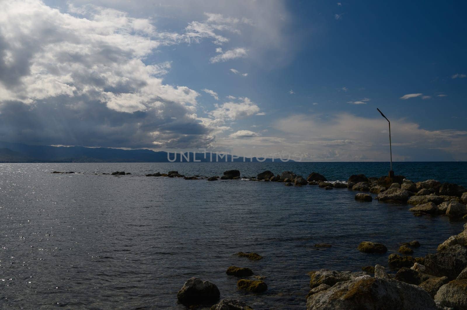 At the edge of a tranquil sea, smooth rocks emerge from the water under a vast sky filled with swirling clouds. The distant mountains create a stunning backdrop, enhancing the serene atmosphere.