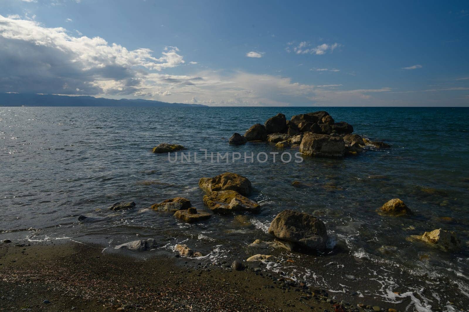 Waves rhythmically embrace the rocky shore, reflecting sunlight on the water's surface. Majestic clouds drift overhead, creating a peaceful and refreshing atmosphere by the ocean.