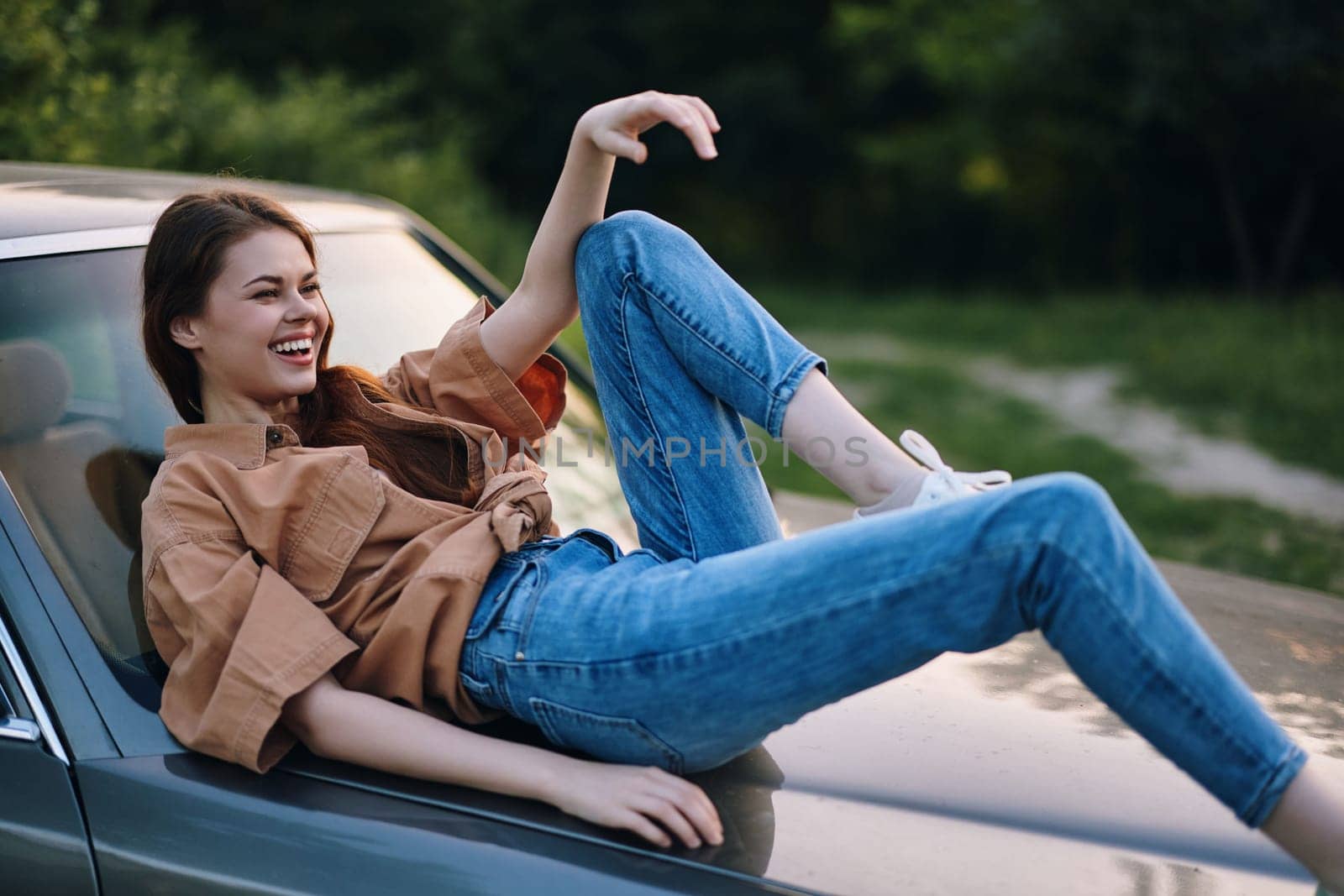 A young woman exudes happiness as she lounges on a vintage car, dressed in casual denim jeans and a trendy brown shirt, set against a vibrant green background, ideal for summer themes.