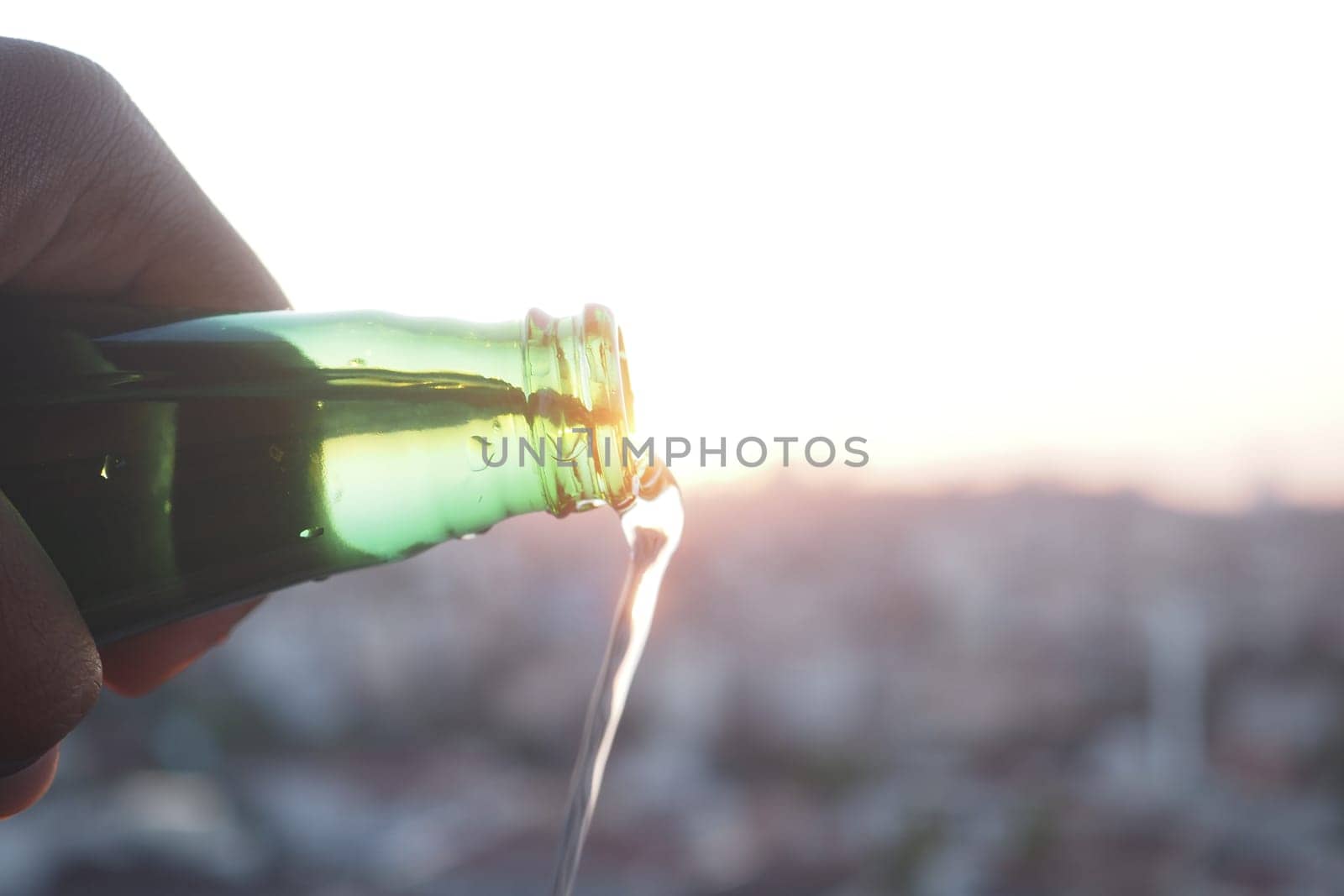 A hand pours a beverage from a green bottle as the sun sets.