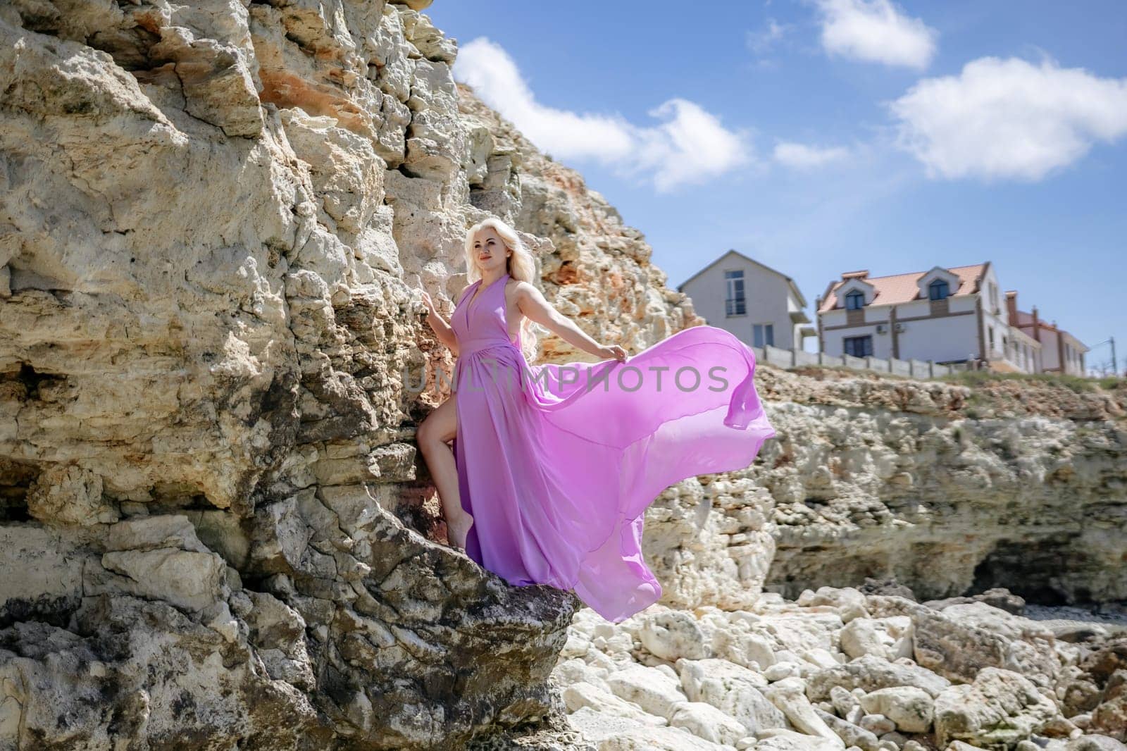 Woman in a flowing pink dress poses dramatically against a rugged cliffside. Houses are seen in the background under a bright sky.