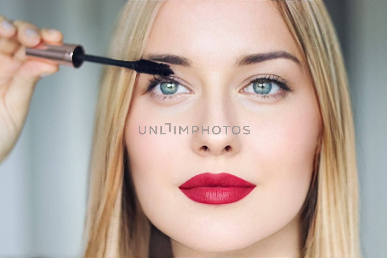 Woman applying mascara with confidence, defined brows and red lipstick. Blonde beauty focused on her eye makeup, cosmetics look