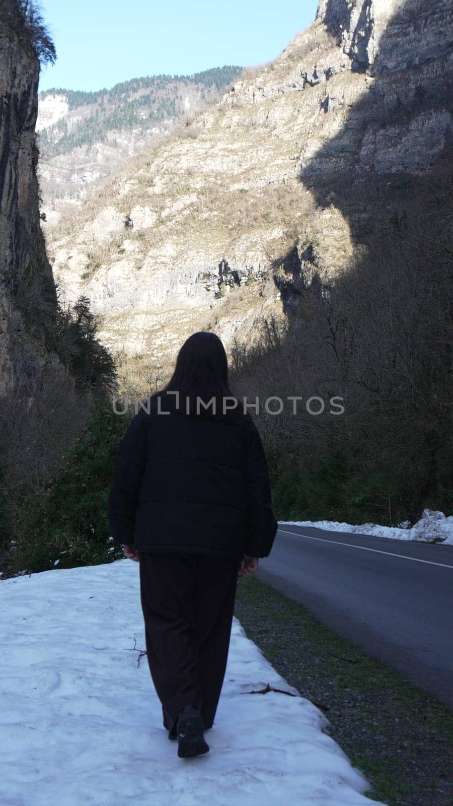 Person in black jacket walks away from camera on snowy path beside road; steep, rocky mountains loom in background.