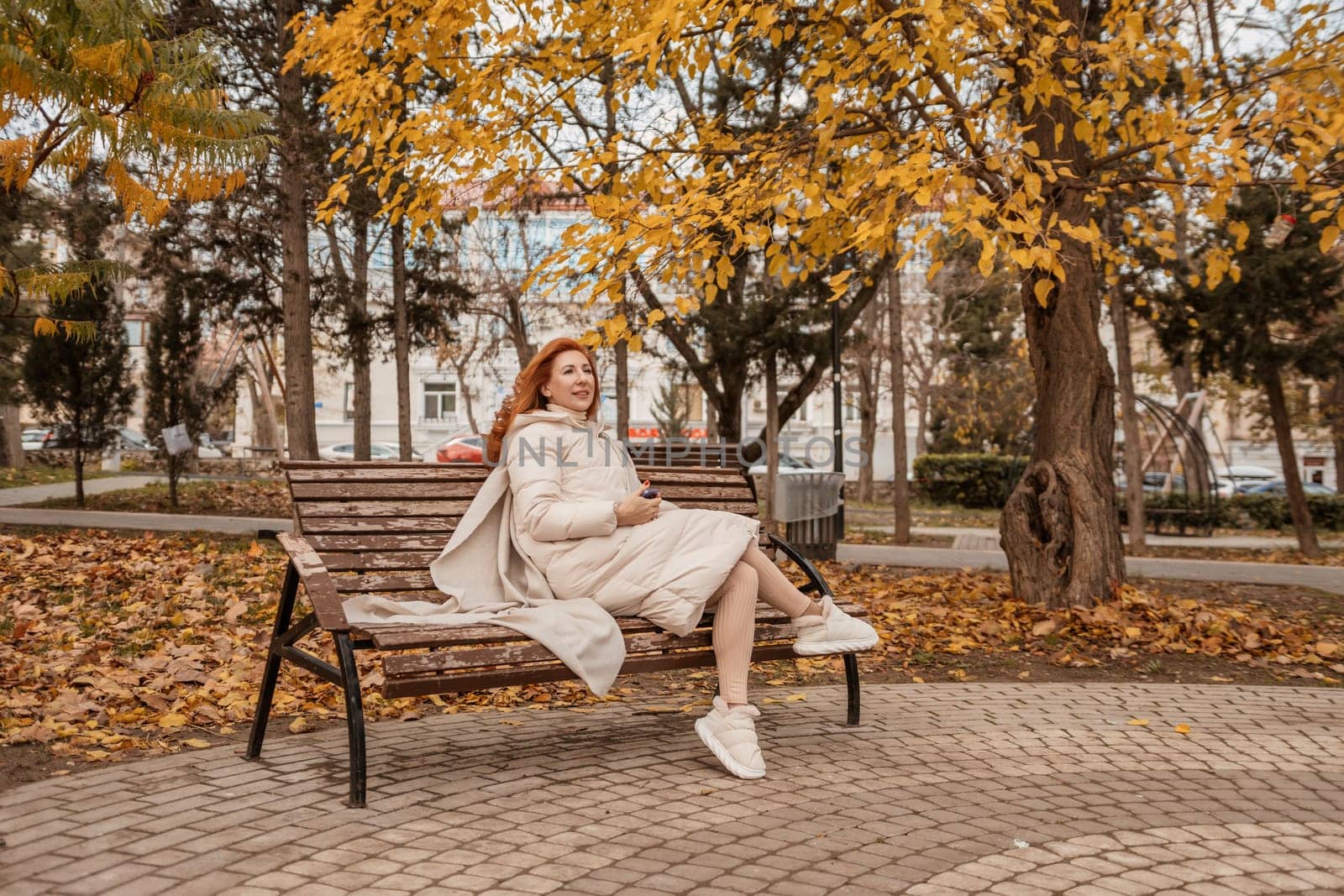 Redhead woman sits on a park bench beneath golden autumn leaves. She wears a cream-colored sweater dress and white sneakers. Fallen leaves cover the ground.