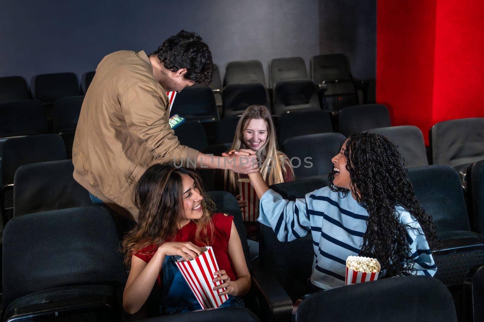 Friends enjoying popcorn and shaking hands while watching an exciting movie in a lively cinema, sharing smiles and laughter together