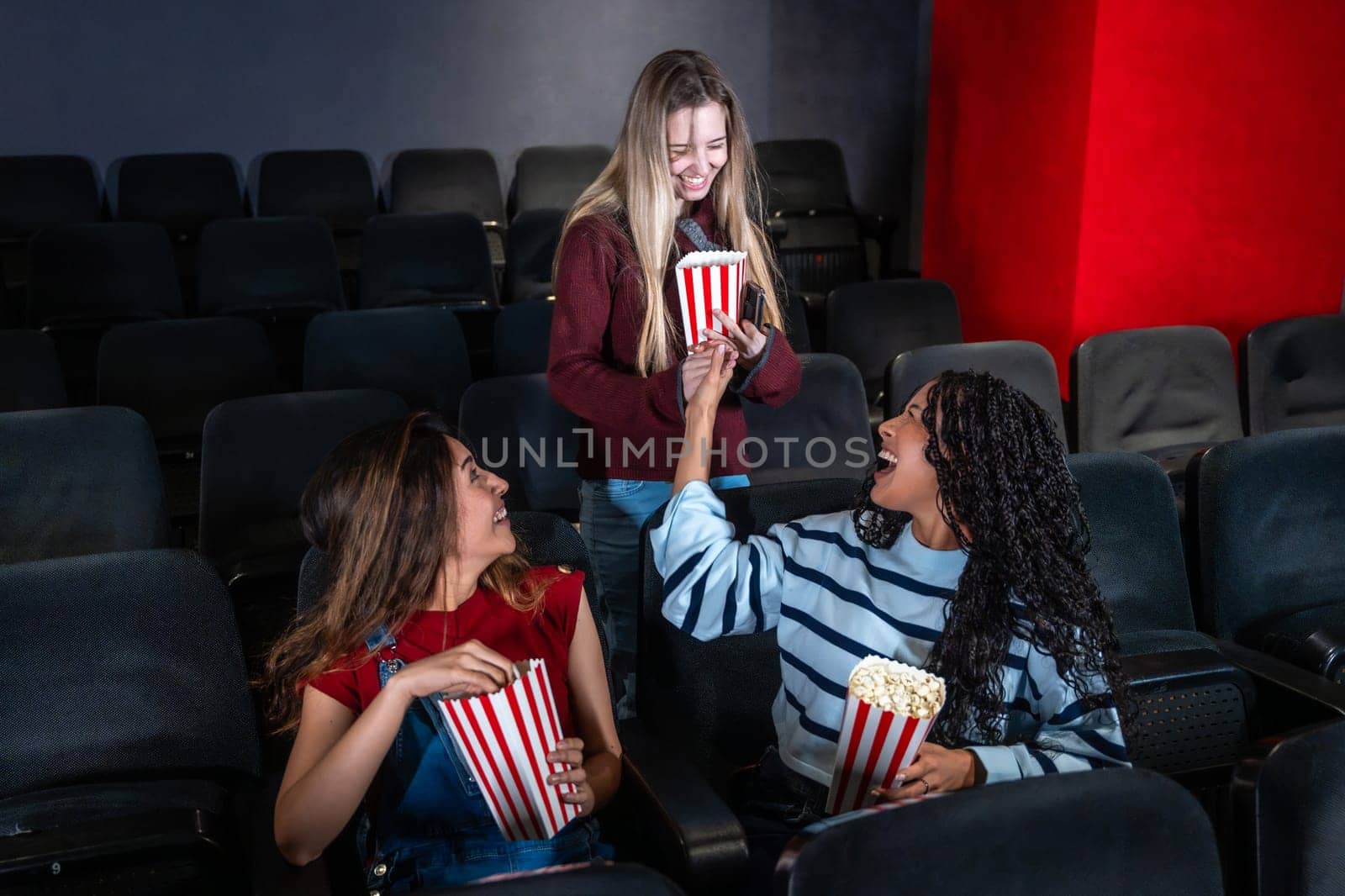 Three female friends are laughing and sharing popcorn while watching a movie in a cinema, enjoying their leisure time together