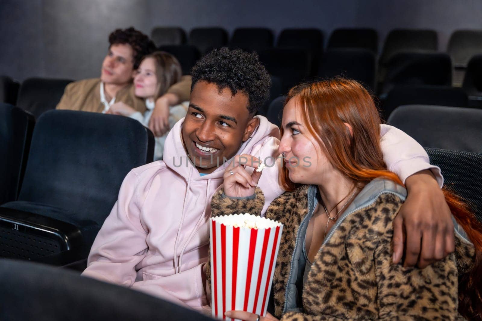 Happy multi ethnic couple sharing popcorn while watching a movie at the cinema, enjoying a date night together