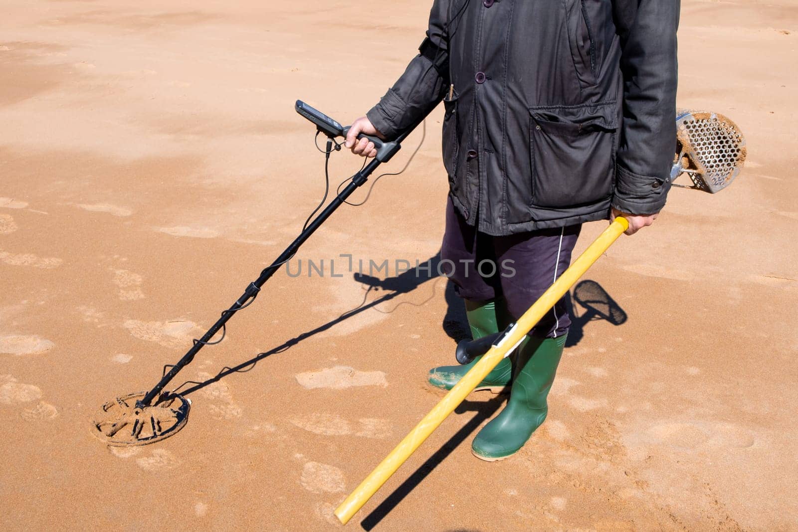 Male adult searching beach for treasures with metal detector and sand scoop.