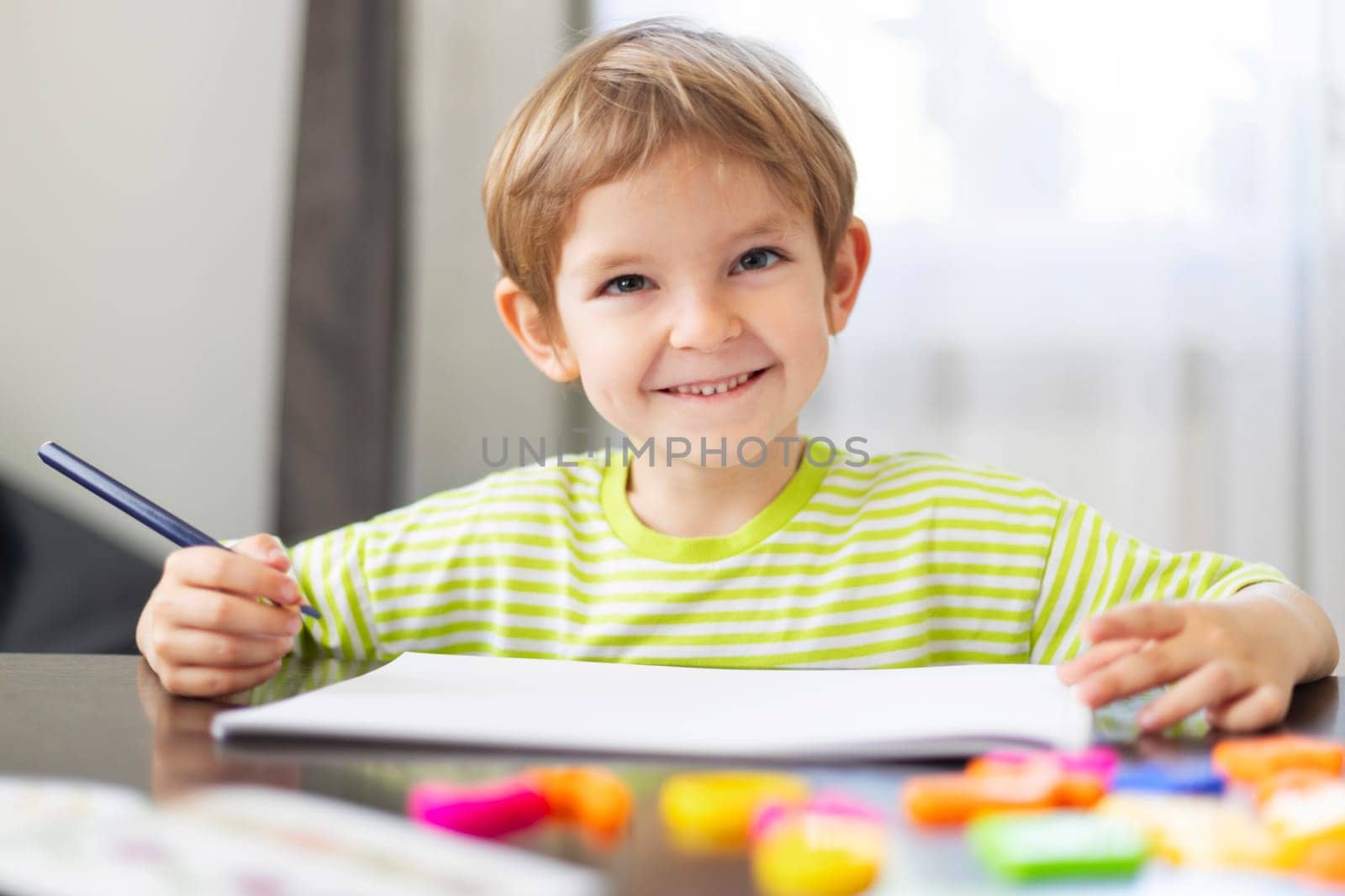 Young boy in a green striped shirt sitting at a table, smiling while holding a pencil and drawing on a white sheet of paper. Colorful educational toys are scattered around