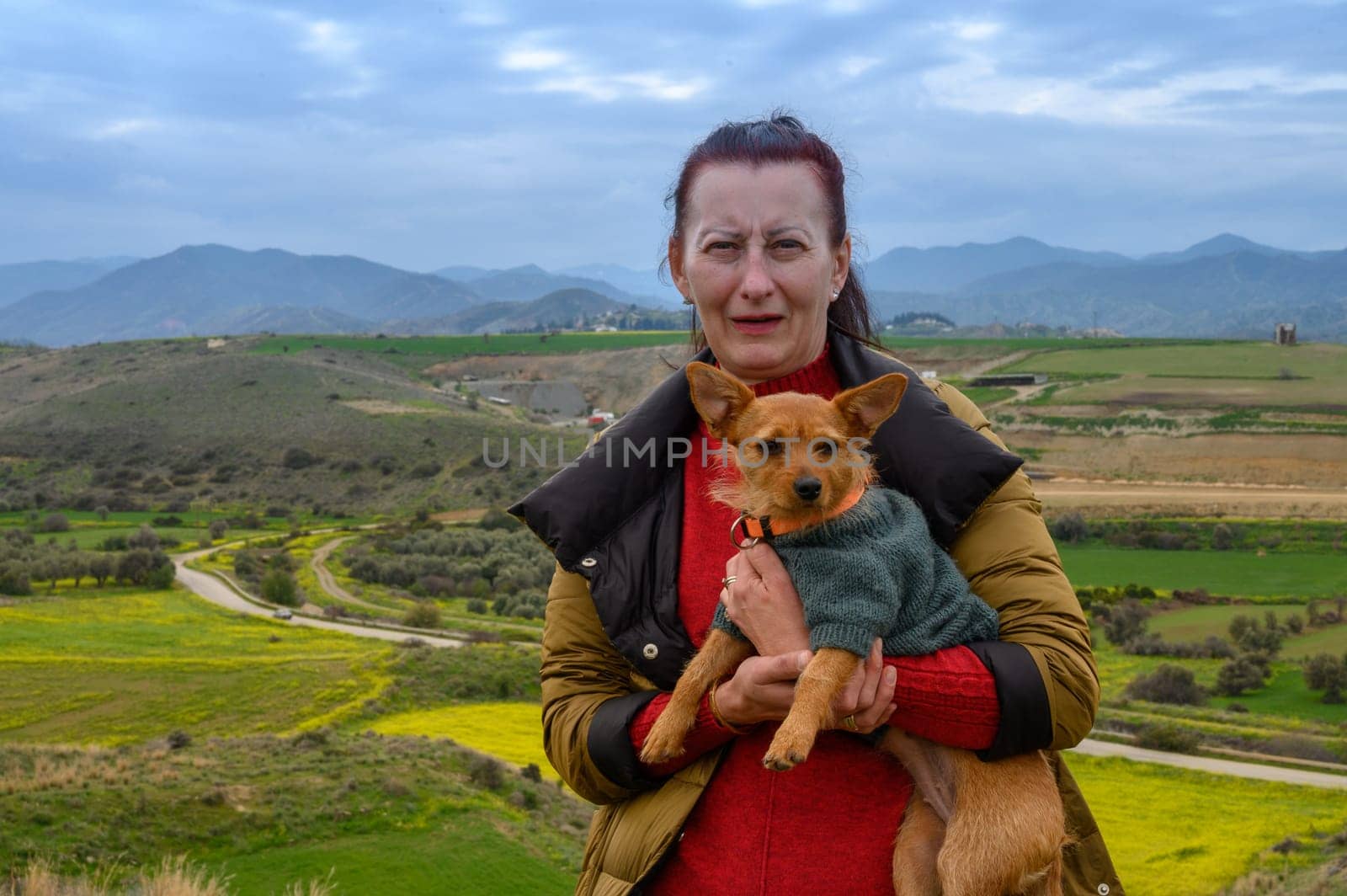 A woman stands on a hilltop, holding her small dog close while surrounded by lush green fields and distant mountains. The crisp air adds to the tranquility of the setting, inviting relaxation.