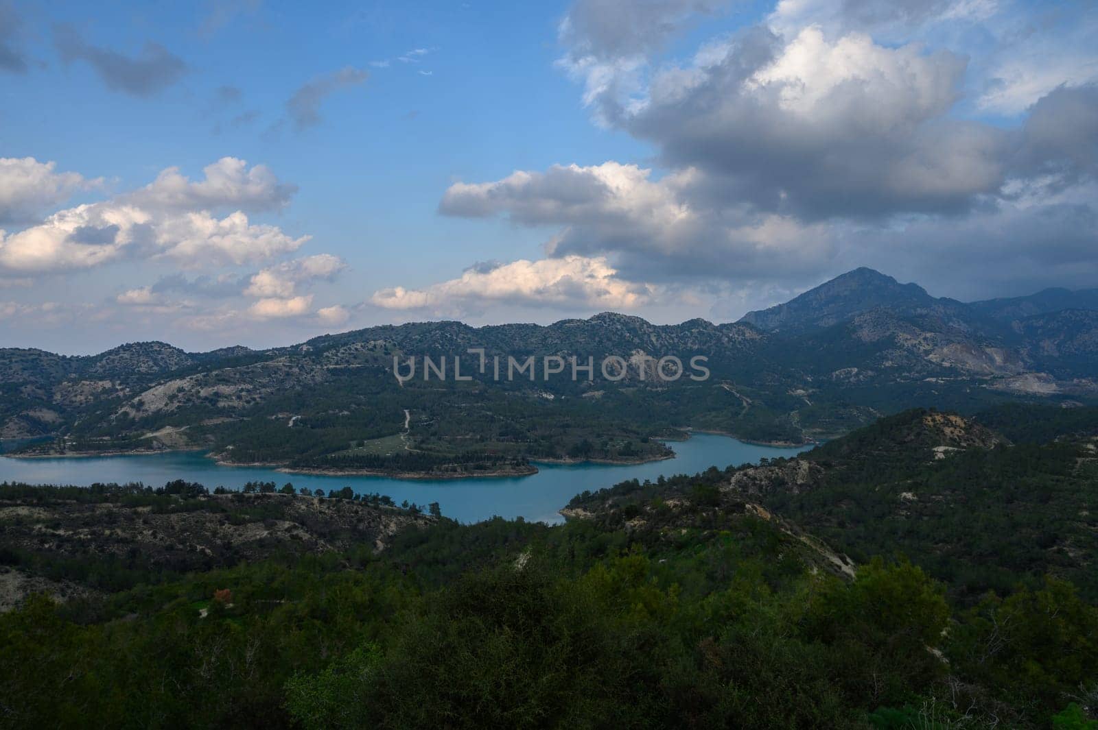 Lush green hills cradle a tranquil turquoise lake, while dramatic clouds drift above mountains at dusk, creating a breathtaking natural panorama.