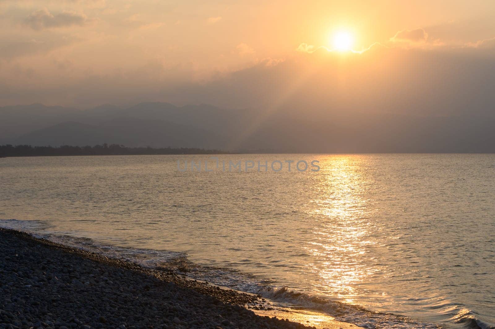 As the sun dips below the horizon, soft golden rays reflect off calm waters, illuminating a pebble-strewn shore. A gentle breeze stirs the peaceful atmosphere at twilight.