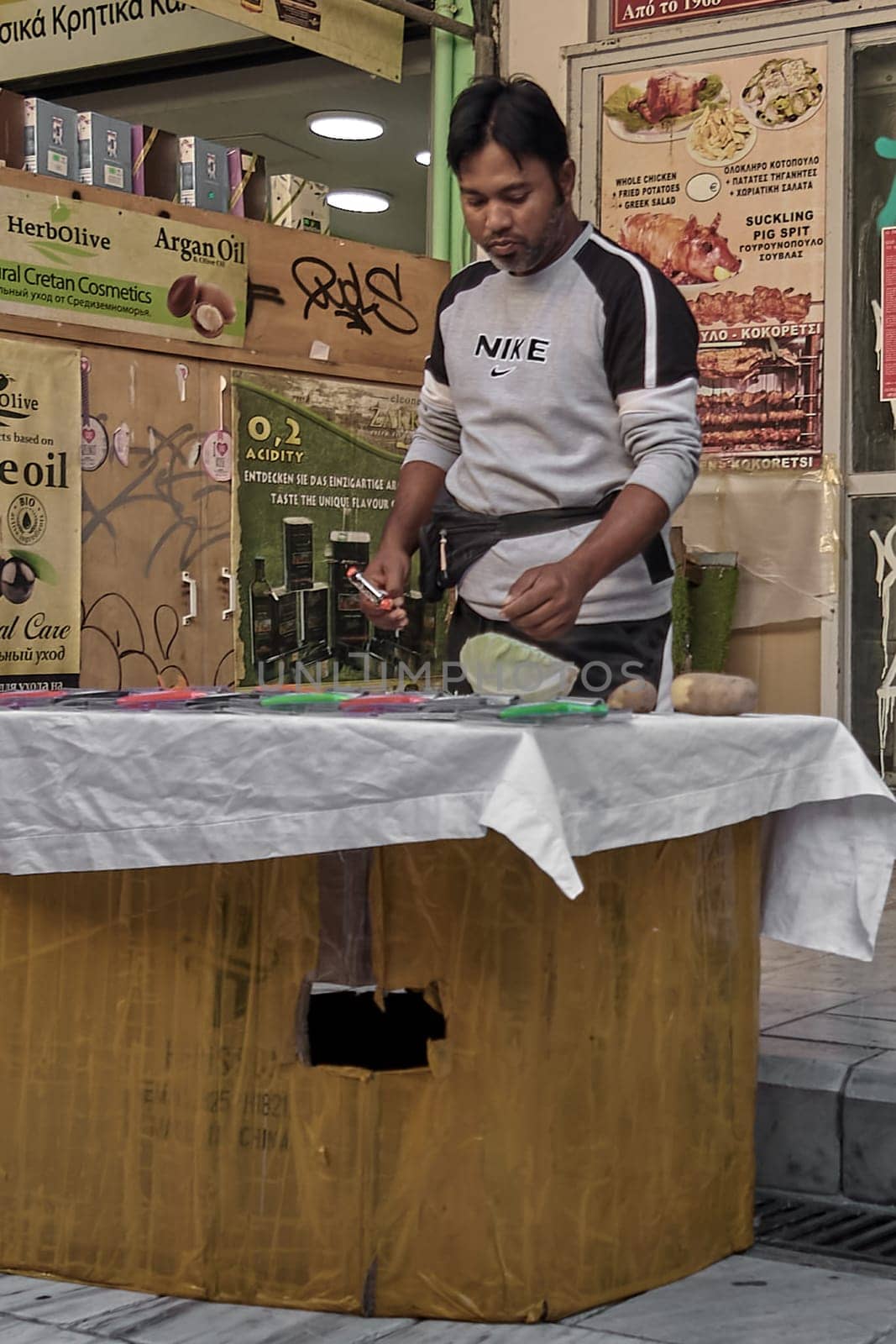 Crete, Greece - April 5, 2025: A lively street vendor shows a cooking utensil amid a bustling market, with logos and promotional materials in the scene.