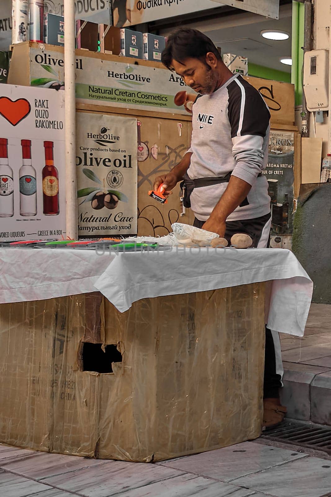 Crete, Greece - April 5, 2025: At a vibrant outdoor market, a merchant demonstrates a kitchen gadget surrounded by fresh produce and advertising posters.