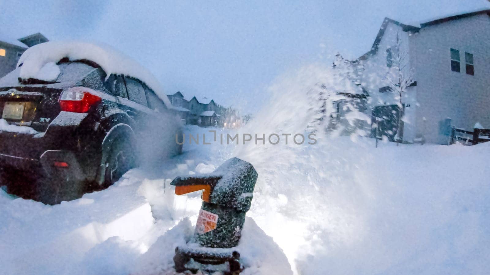 Denver, Colorado, USA-February 4, 2024-Snow blower is at work in the early evening light, clearing a path in a neighborhood where houses and a large snow pile are softly illuminated against the dusky sky.