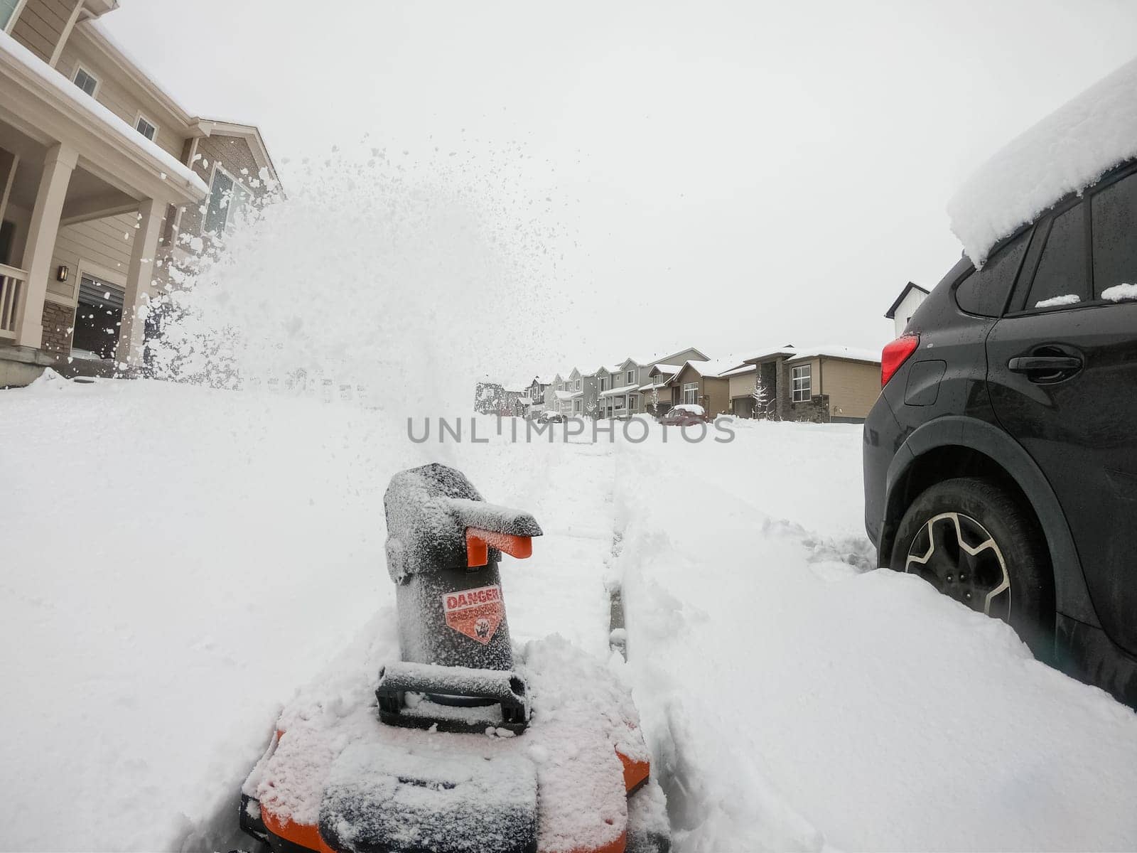 Denver, Colorado, USA-February 4, 2024-View from a snow-blanketed driveway captures a residential area mid-snowfall, with houses partially obscured by the cascading white flakes and a snow-covered car in the foreground.