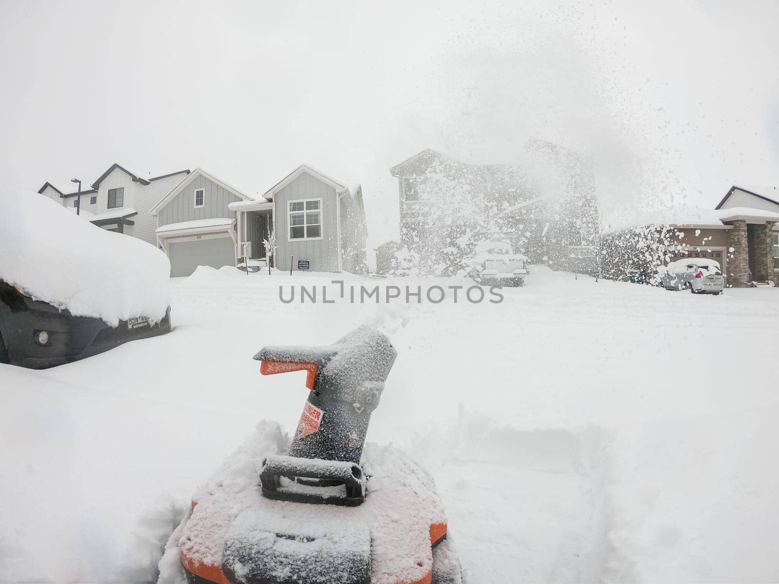 Denver, Colorado, USA-February 4, 2024-View from a snow-blanketed driveway captures a residential area mid-snowfall, with houses partially obscured by the cascading white flakes and a snow-covered car in the foreground.