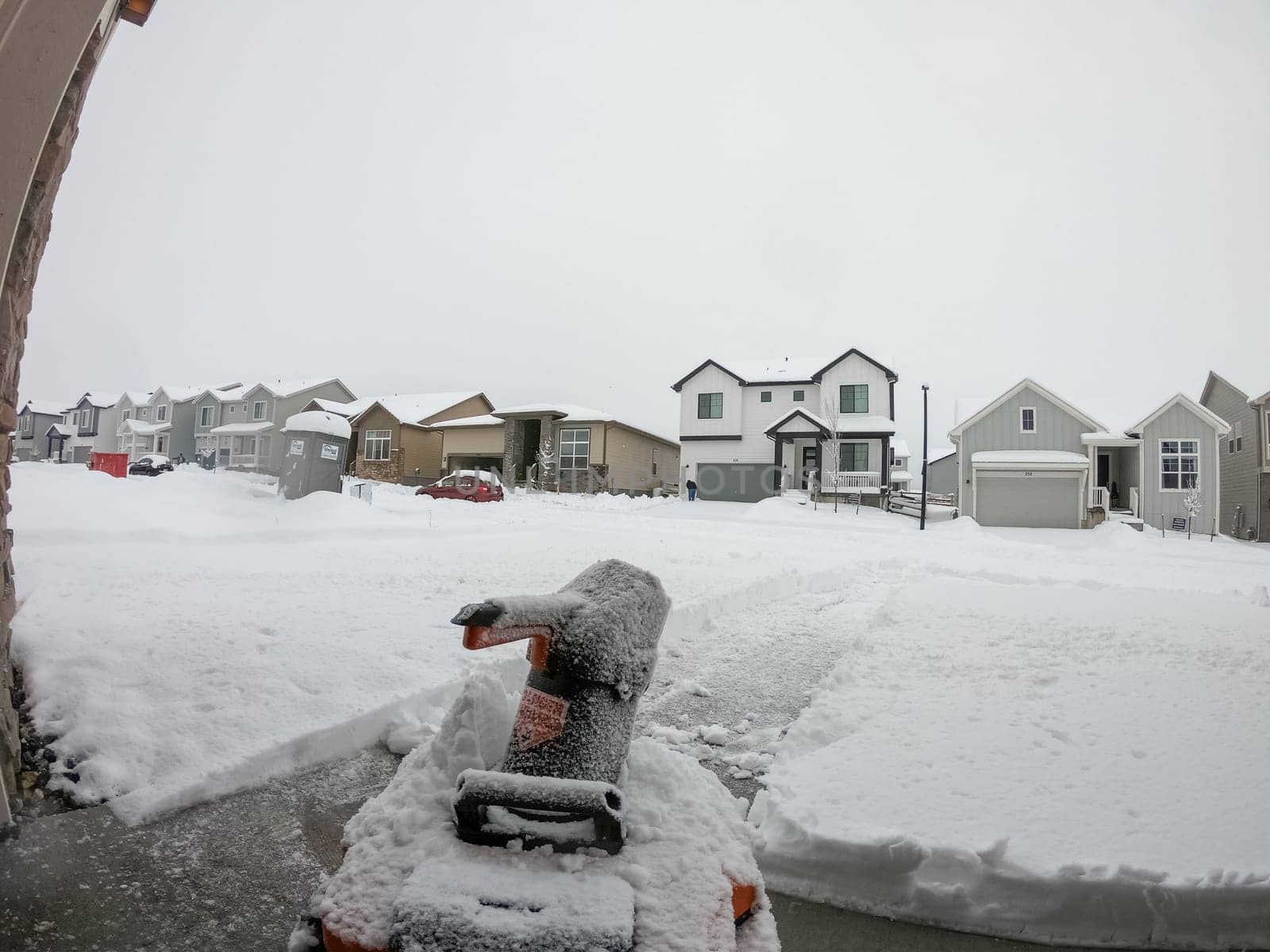 Denver, Colorado, USA-February 4, 2024-View from a snow-blanketed driveway captures a residential area mid-snowfall, with houses partially obscured by the cascading white flakes and a snow-covered car in the foreground.