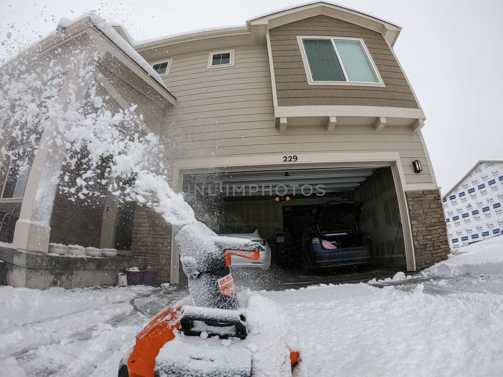 Denver, Colorado, USA-February 4, 2024-View from a snow-blanketed driveway captures a residential area mid-snowfall, with houses partially obscured by the cascading white flakes and a snow-covered car in the foreground.