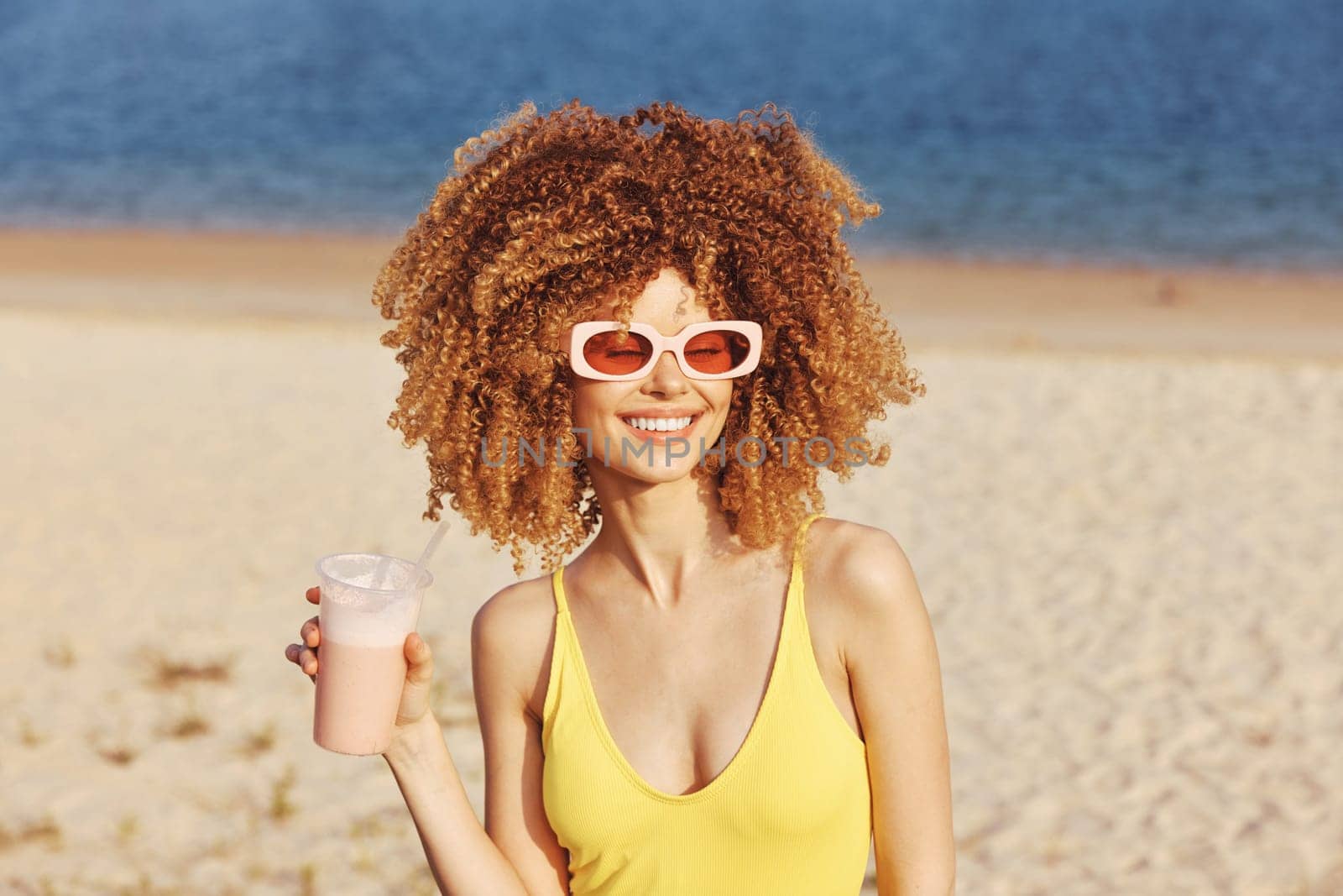Cheerful woman with curly hair wearing sunglasses and a yellow swimsuit enjoys a refreshing drink by the seaside