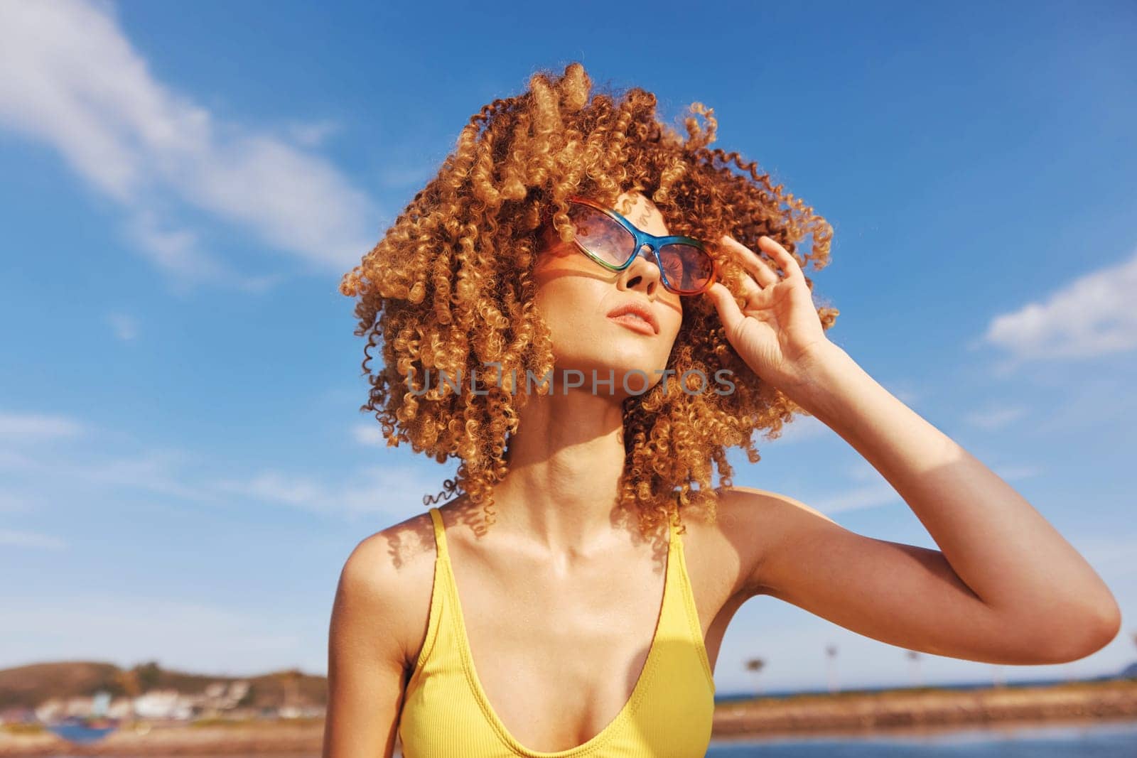 A vibrant scene of a cheerful woman with curly hair, dressed in a yellow swimsuit, relaxing on a sunny beach with clear blue skies and soft clouds