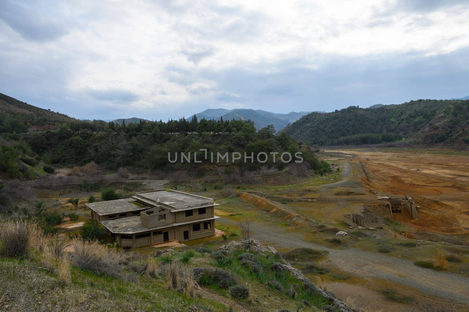 A derelict building stands isolated amidst a dry landscape, surrounded by rolling hills and a cloudy sky, evoking a sense of eerie abandonment and faded memories.