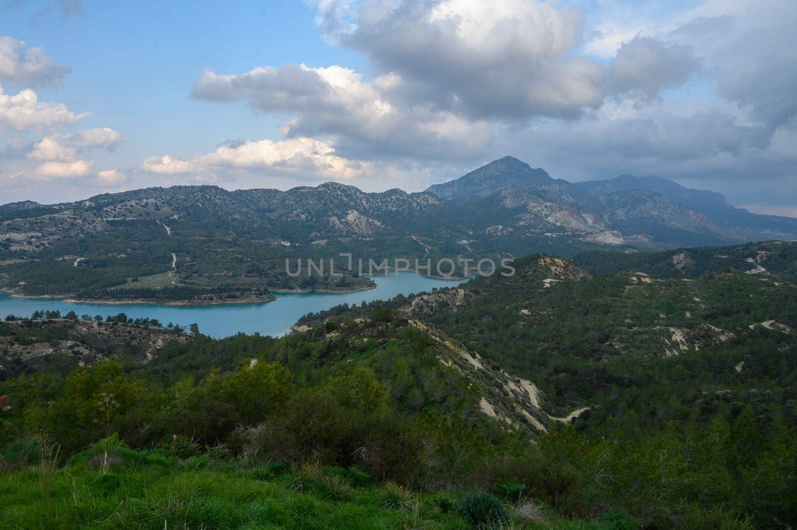 Bright clouds float above a serene lake surrounded by verdant hills and majestic mountains under a soft afternoon light, creating a peaceful atmosphere in nature.