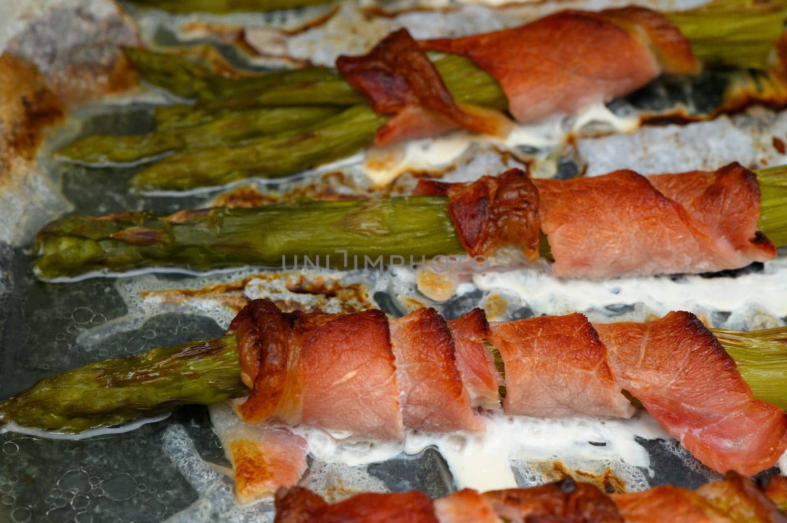 Fresh asparagus spears are delightfully wrapped in crisp bacon, sizzling and browning on a baking sheet. The warmth of the kitchen adds to the inviting aroma of this delicious dish.