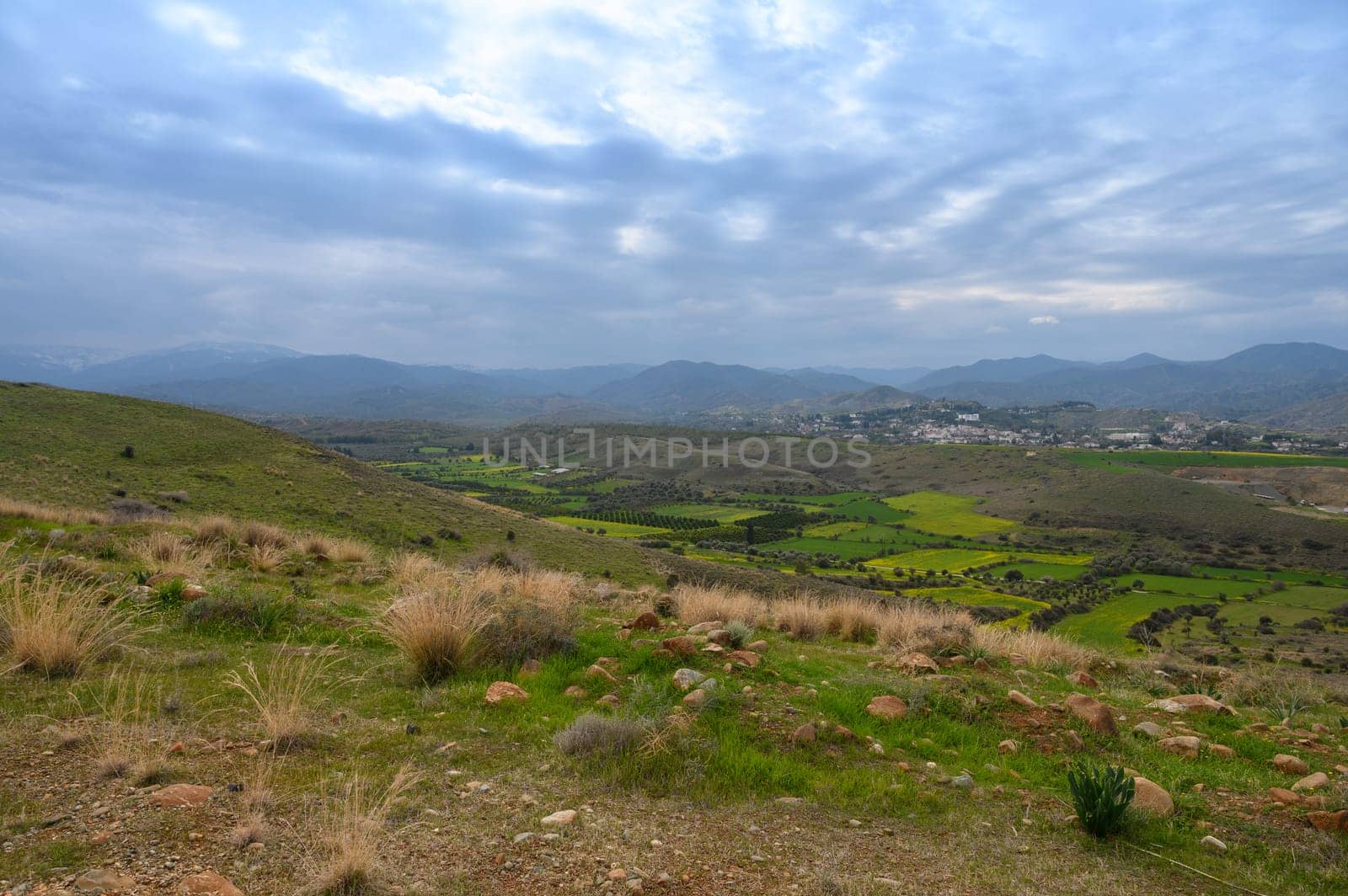 Rolling hills cradle a peaceful valley adorned with patches of lush greenery and distant villages, illuminated by a soft, muted light. Nature's quietude prevails in this serene haven.
