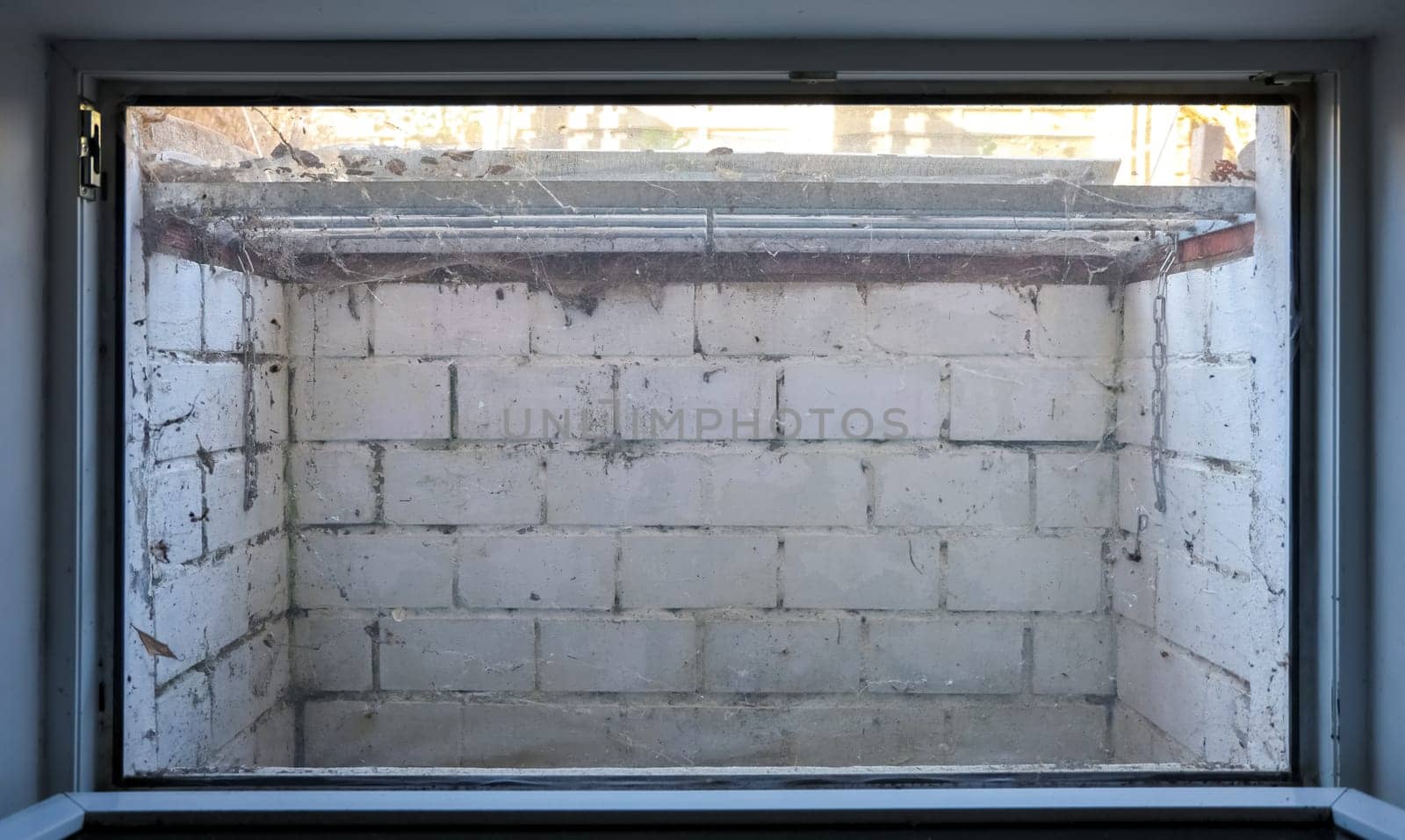 View through a window showing a small, dirty basement with white brick walls, a rusty metal grate, and spiderwebs, suggesting neglect and disrepair