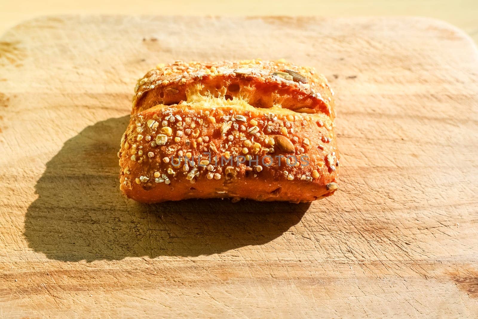Small, appetizing multigrain bun sits on a worn wooden cutting board, casting a subtle shadow in the warm sunlight