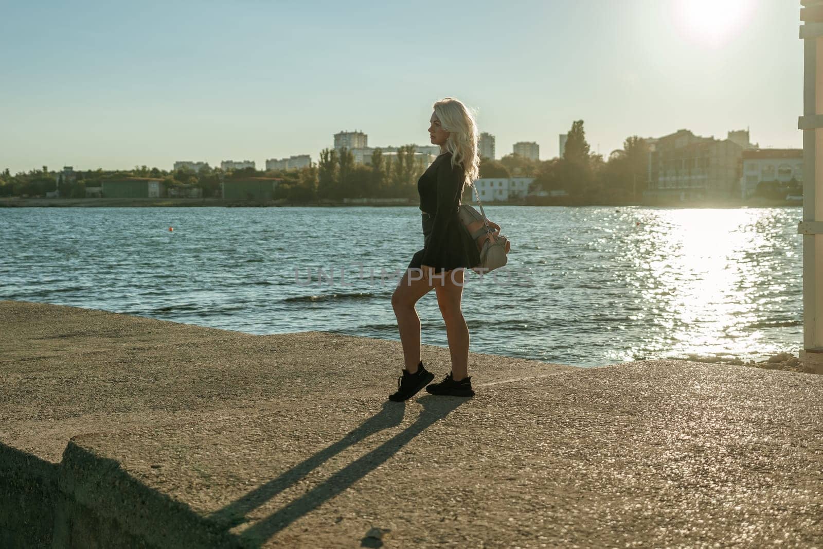 A woman in a black dress stands on a pier overlooking a body of water, with a city skyline in the background. The sun is setting, casting a warm glow on the scene.