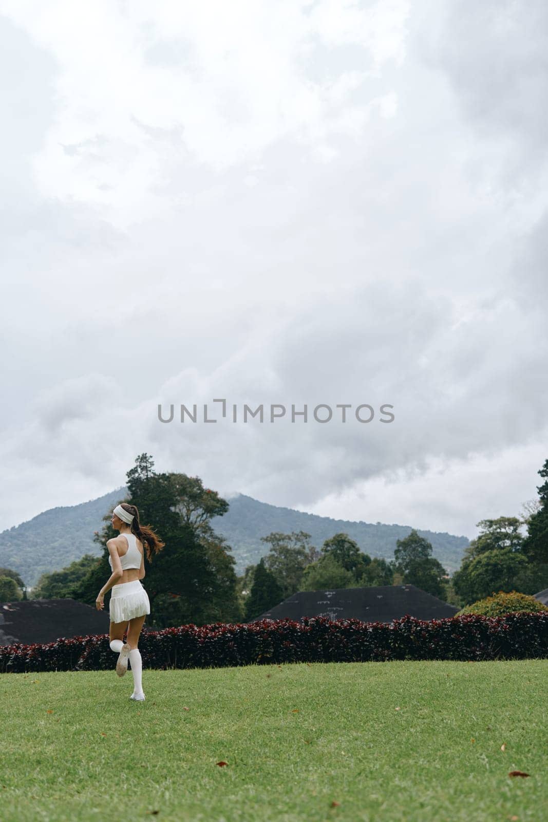 A joyful young woman in white athletic attire runs across a vibrant green lawn, with a stunning mountain landscape in the background, showcasing an active and fresh lifestyle